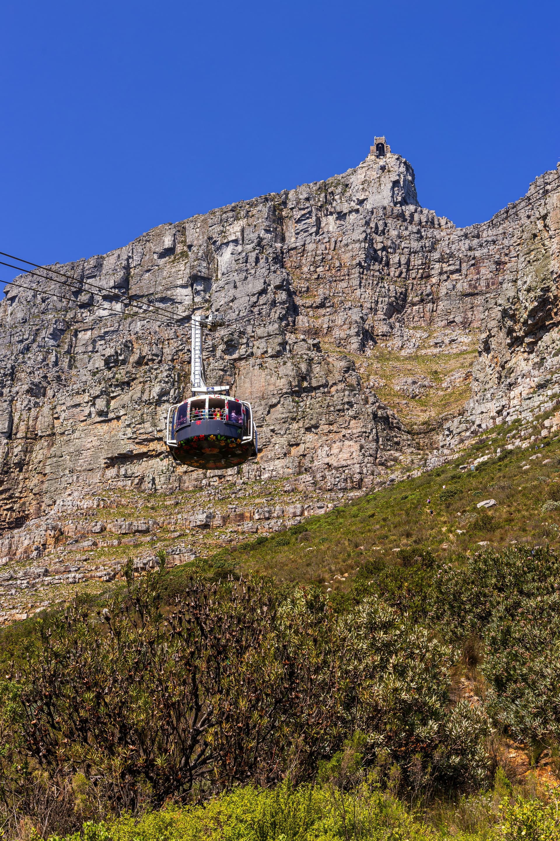 🏞️ Cable Car on Table Mountain – Cape Town, South Africa A cable car hangs midair against the steep cliffs of Table Mountain, gliding upward on taut lines toward the summit. Rocky textures and sparse vegetation blanket the mountain’s rugged surface, while soft clouds float overhead. Below, the faint outlines of Cape Town peek through, adding depth to this high-altitude adventure above one of South Africa’s most iconic natural landmarks.